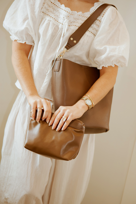 A woman holding a brown Modern+Chic vegan leather Sophie Bucket Bag and coordinating zipper pouch for Women – with a blurred background.