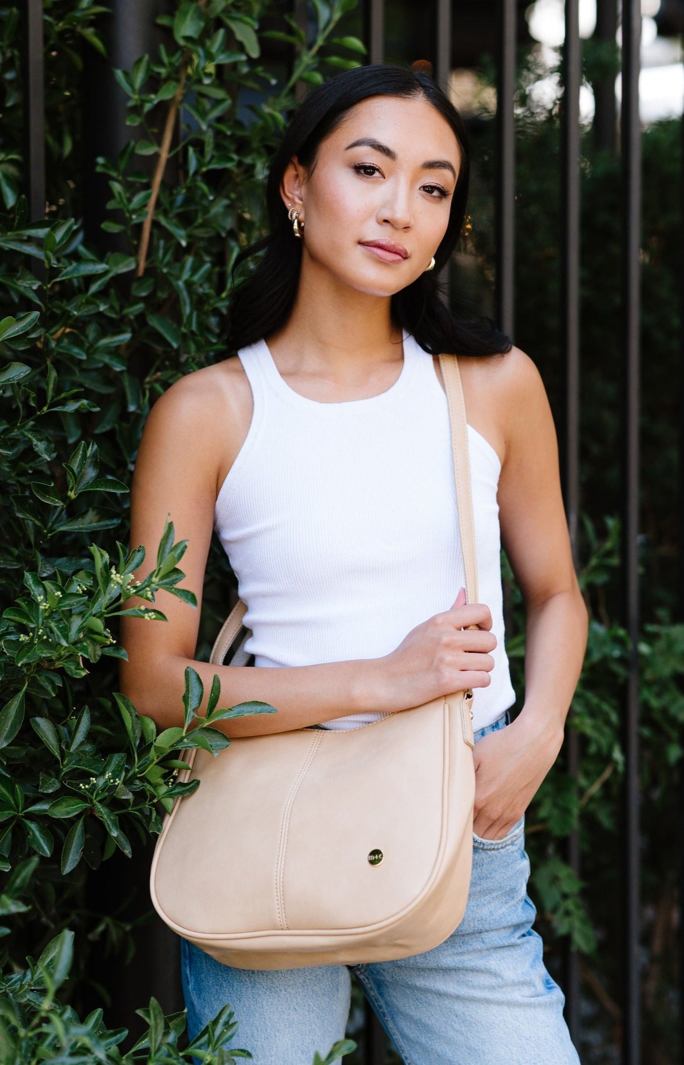 A woman standing in front of greenery and a fence wearing a cream crossbody.