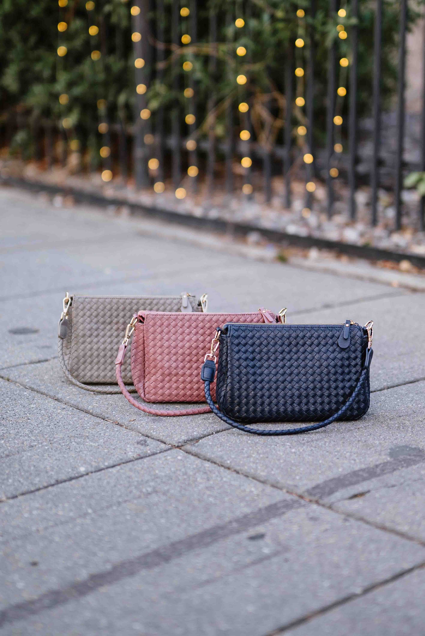 Three woven Modern + Chic Elle Woven Crossbody Bag in gray, pink, and blue on a paved surface with blurred lights in the background.