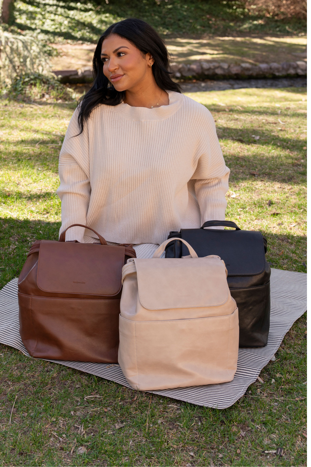 A woman kneeling on a black and white striped blanket sitting on the grass with three different Modern+Chic Brielle 2.0 Vegan Leather Convertible Backpack Purses for Women sitting in front of her in the  shades dark brown, taupe and black.