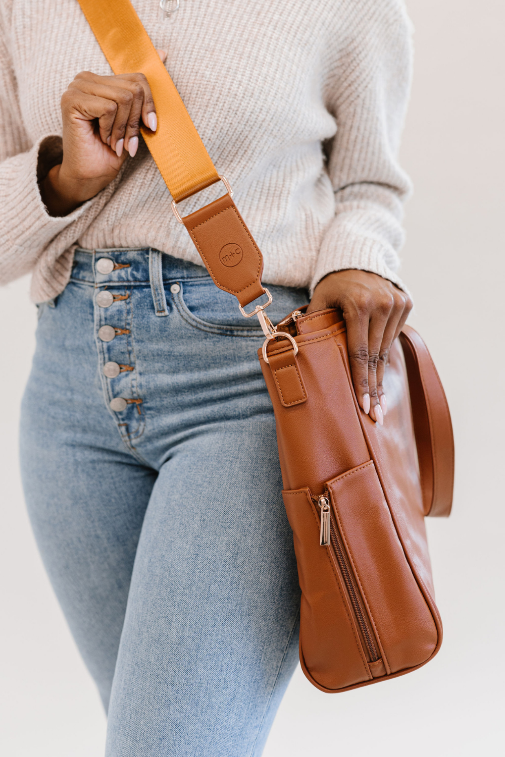 The strap on the brown tote has a stamped circular m+c logo on it. This tote also features side zipper pockets.
