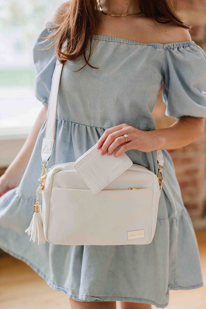 Women posing with a white Modern + Chic Alice Crossbody vegan leather bag with gold hardware, displaying a removable tassel, holding a matching wallet in her hand. 