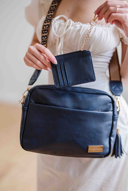 Women posing with a navy Modern + Chic Alice Crossbody vegan leather bag with gold hardware, matching key leash and wallet with a removable tassel attached.