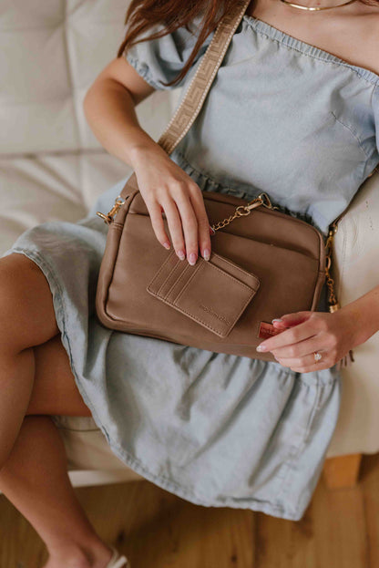 A red-haired woman modeling a Fawn colored Modern + Chic Alice Crossbody bag, showing the included key leash and wallet. 