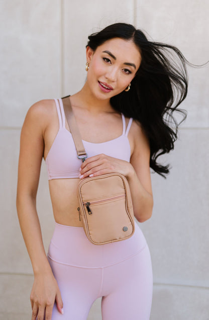 A woman wearing a pink sports bra and leggings is showcasing a pink Lainey Convertible Crossbody bag with silver hardware, attached to her shoulder via an adjustable strap.