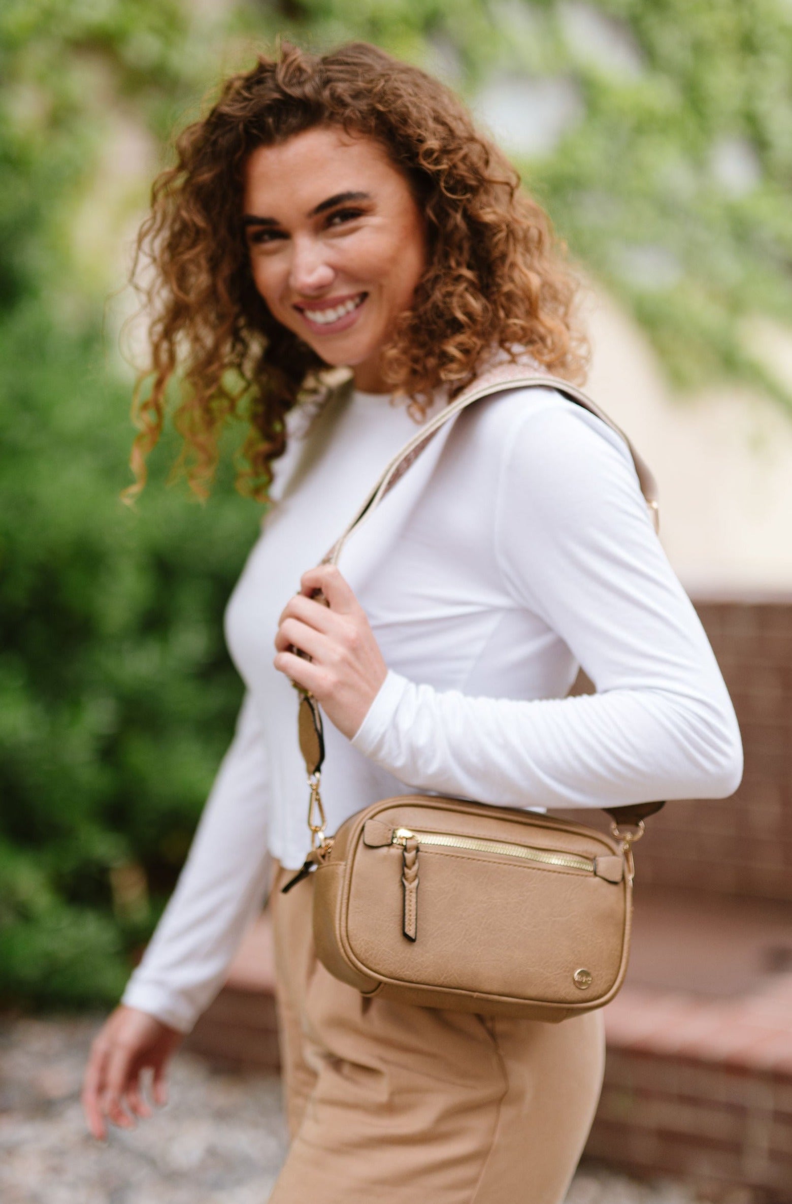 Woman wearing white shirt, tan pants and a Modern + Chic Gentry Vegan Leather Crossbody Bag in tan.