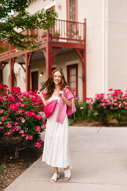 A woman wearing a white dress and pink sweater draped over her shoulders with a hot pink oversized Modern+Chic Presley Oversized Sling Bag for Women with gold hardware across her torso.