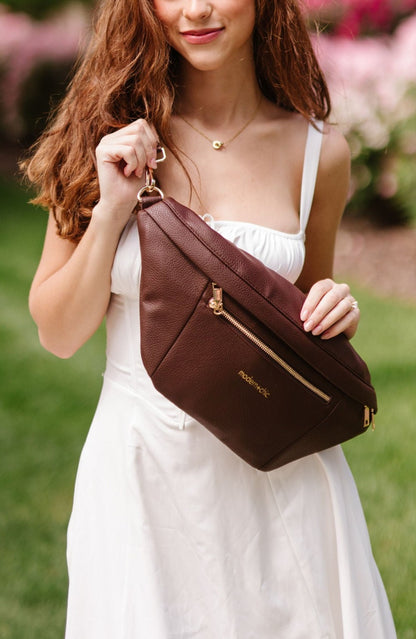 A woman wearing a white dress holding an oversized brown Modern+Chic Presley Oversized Sling Bag for Women with gold hardware as she wears it crossbody.