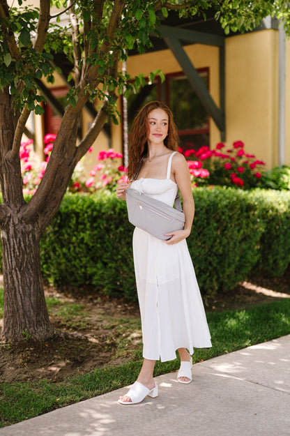 A woman wearing a white dress and an oversized grey Modern+Chic Presley Oversized Sling Bag for Women with gold hardware across her torso.