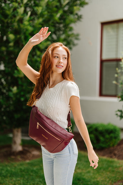 A woman wearing a white cropped sweater and an oversized wine colored Modern+Chic Presley Oversized Sling Bag for Women with gold hardware across her torso.