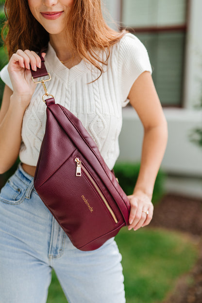 A woman wearing a white cropped sweater and an oversized wine colored Modern+Chic Presley Oversized Sling Bag for Women with gold hardware across her torso.