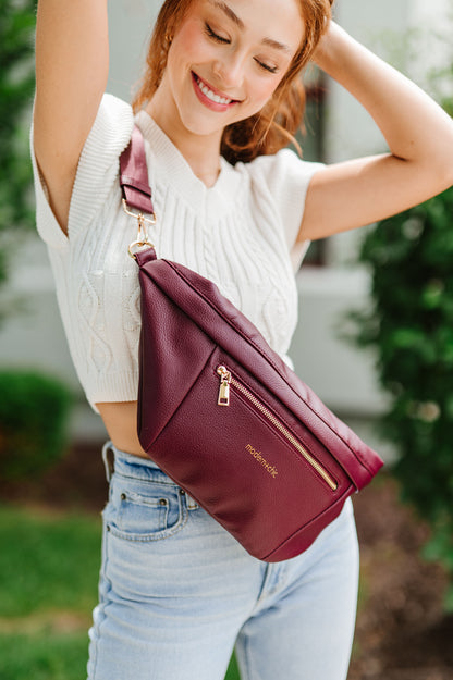 A woman wearing a white cropped sweater and an oversized wine colored Modern+Chic Presley Oversized Sling Bag for Women with gold hardware across her torso.