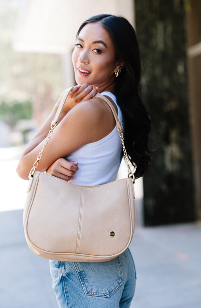 A woman wearing a cream Modern + Chic Marissa Vegan Leather Crossbody handbag over her shoulder displaying the gold chain accents on the strap.