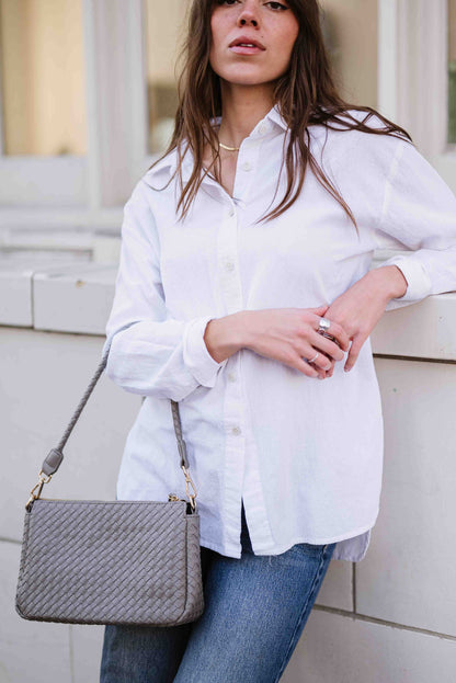 Woman wearing a white shirt and jeans holding a gray Modern + Chic Elle Woven Crossbody Bag for Women against a light-colored building.