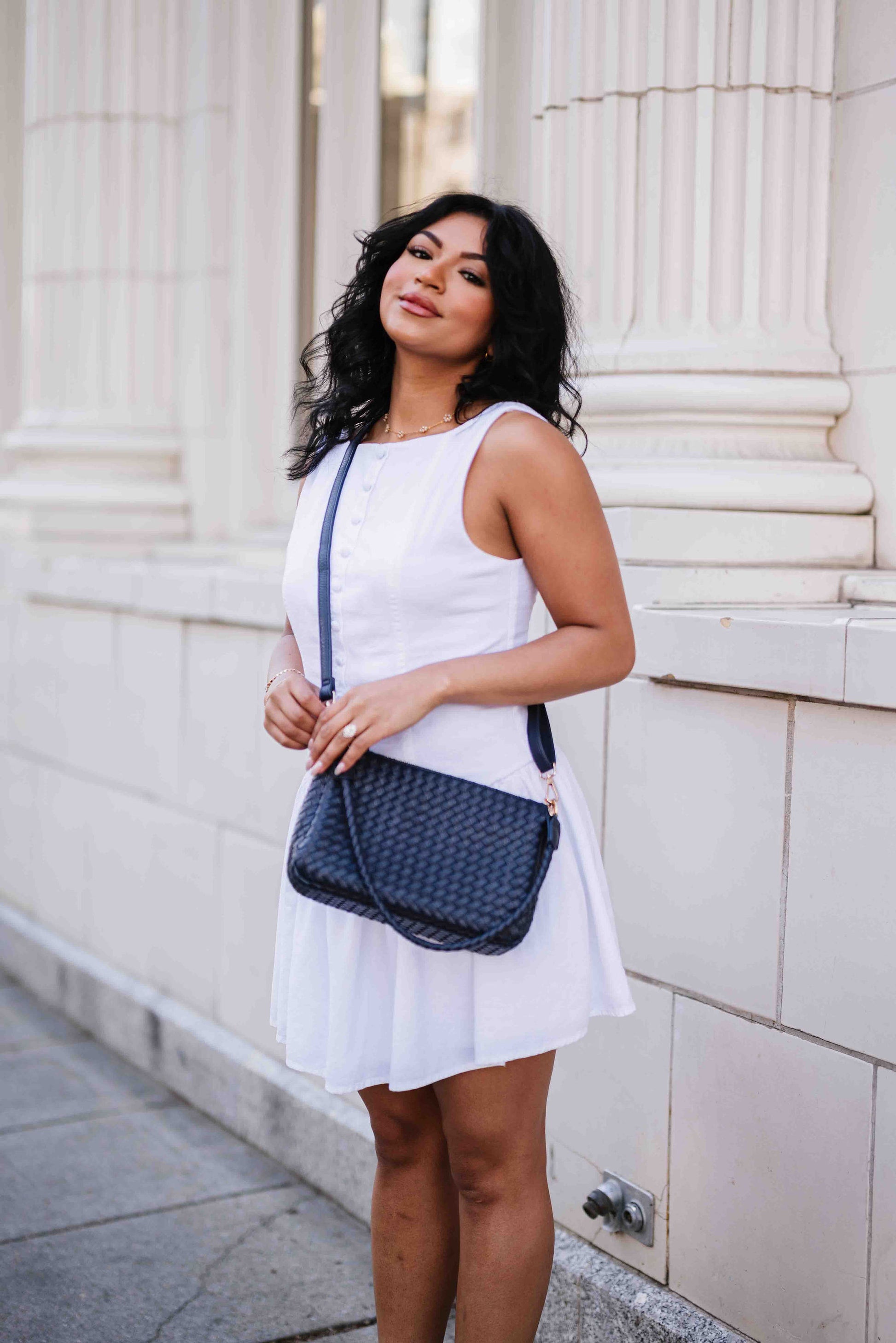Woman in a white dress holding a blue A woman wearing a Modern + Chic Elle Woven Crossbody Bag for Women over crossbody outdoors against a light-colored building.