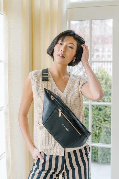 A woman wearing a cream sweater vest and an oversized black Modern+Chic Presley Oversized Sling Bag for Women with gold hardware across her torso.
