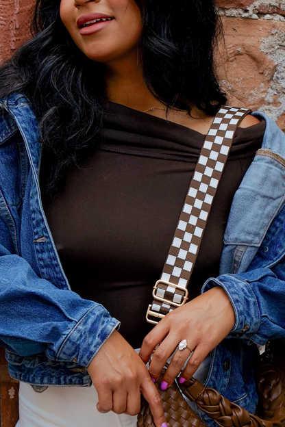 Woman wearing a denim jacket and brown top with a brown and white Modern+Chic Lizzie checkered adjustable strap bag against a brick wall.