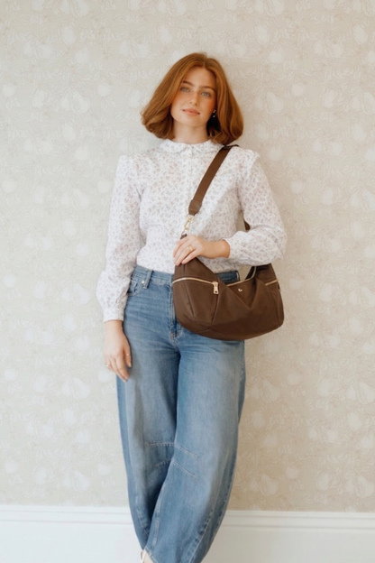Woman holding a brown Modern + Chic Parker Crescent Crossbody Bag for Women against a light wall.