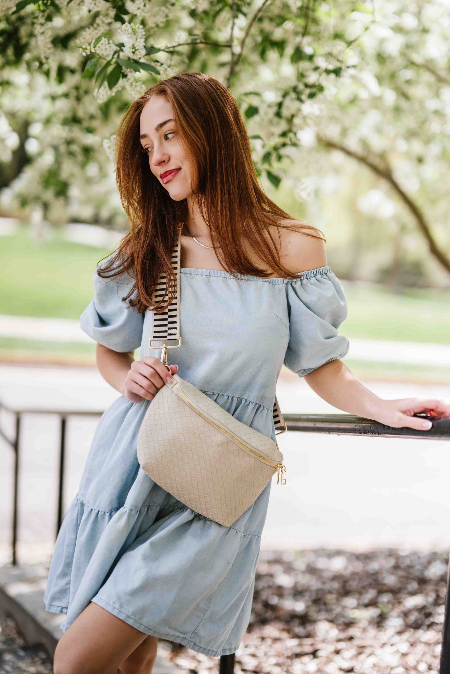 Woman wearing an off the shoulder blue dress and a cream criss cross style Modern + Chic Rory Oversized Belt Bag for Women with a gold zipper and hardware crossbody.