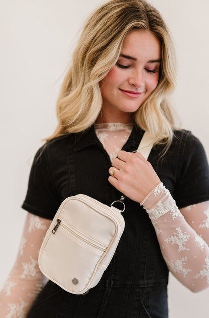 A woman wearing a lace long sleeve top under a black jumpsuit and a white Modern + Chic Lainey Convertible Crossbody Bag for Women crossbody.