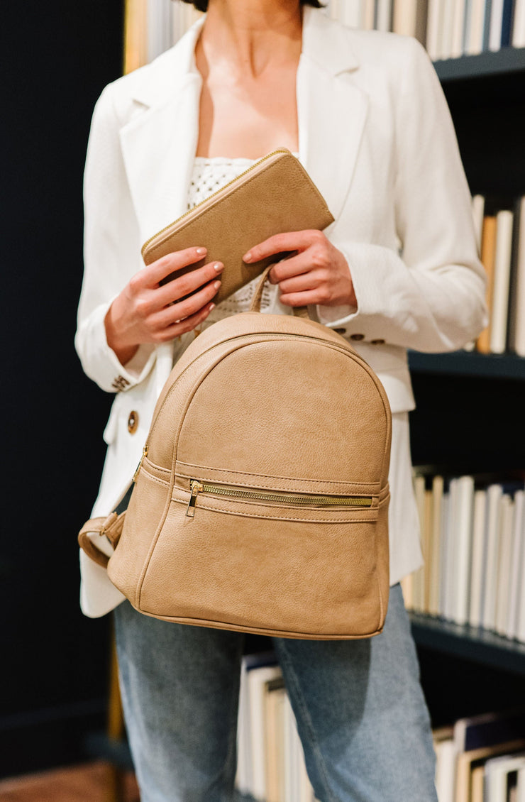Woman standing in a library holding a taupe dome backpack with a matching wallet.