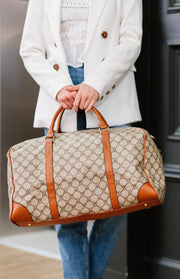 Woman holding a travel bag that is beige and dark brown with camel trim and gold hardware.